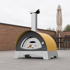 Alfa Ciao wood fired oven in yellow on paving beside a brick wall, a parasol and chairs behind