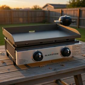 Blackstone 22-inch tabletop griddle on a wooden deck at sunset, stainless front plate and two control knobs, seen from the front left
