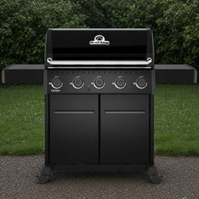 Black Broil King Crown 520 Shadow gas grill on gravel, lid closed, five knobs above two cabinet doors, hedge behind