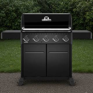 Black Broil King Crown 520 Shadow gas grill on gravel, lid closed, five knobs above two cabinet doors, hedge behind