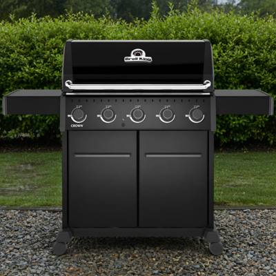 Black Broil King Crown 520 Shadow gas grill on gravel, lid closed, five knobs above two cabinet doors, hedge behind