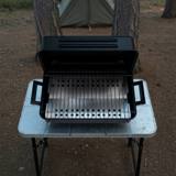 Black Char-Broil portable charcoal grill open on a camp table, pine trees and a tent behind it