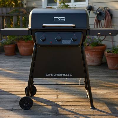 Black Char-Broil 3-burner gas grill on a weathered deck, lid closed, both side shelves out, potted plants behind