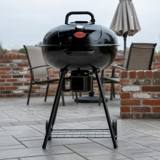 Black Char-Griller 22-inch kettle grill on gray patio paving beside a brick wall, lid closed, ash bucket under the bowl