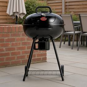 Black Char-Griller 22-inch kettle grill on gray patio paving beside a brick wall, lid closed, ash bucket under the bowl