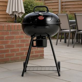 Black Char-Griller 22-inch kettle grill on gray patio paving beside a brick wall, lid closed, ash bucket under the bowl