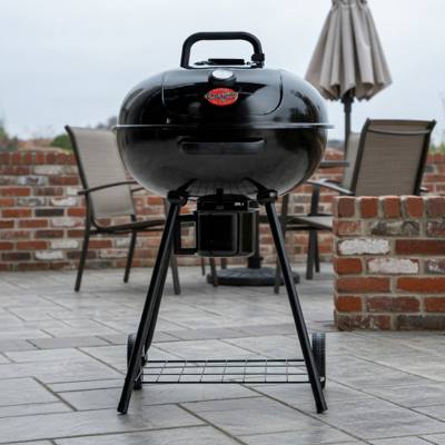 Black Char-Griller 22-inch kettle grill on gray patio paving beside a brick wall, lid closed, ash bucket under the bowl