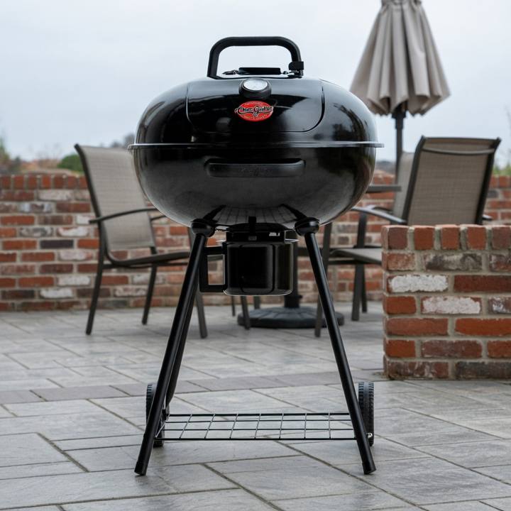 Black Char-Griller 22-inch kettle grill on gray patio paving beside a brick wall, lid closed, ash bucket under the bowl