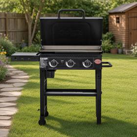Black Char-Griller 3-burner flat top on a mown lawn, lid raised over bare steel plate, three knobs, shelf folded out