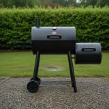 Black Char-Griller Smokin' Pro barrel smoker on wet gravel, side fire box at right, smokestack up, hedge behind