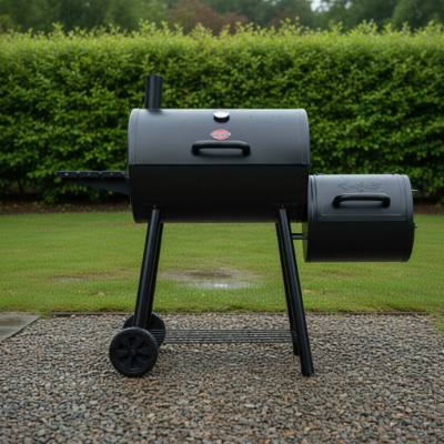 Black Char-Griller Smokin' Pro barrel smoker on wet gravel, side fire box at right, smokestack up, hedge behind