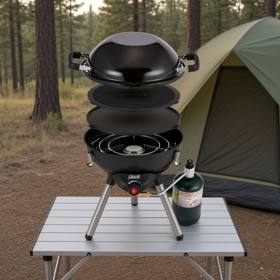 Coleman 4-in-1 cooking system stacked on a camp table, wok, grill plate and burner visible, propane cylinder alongside and a tent behind