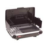 Coleman 2209294 tabletop grill and stove open on a picnic table at a wooded campsite, grate beside the burner