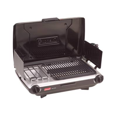 Coleman 2209294 tabletop grill and stove open on a picnic table at a wooded campsite, grate beside the burner