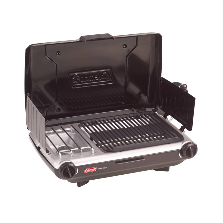 Coleman 2209294 tabletop grill and stove open on a picnic table at a wooded campsite, grate beside the burner