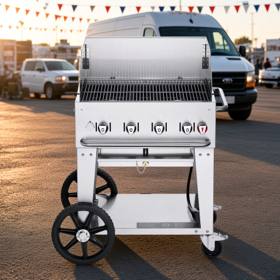 Crown Verity 30 inch mobile grill in stainless steel, windguard panels raised, parked on asphalt beside a white van