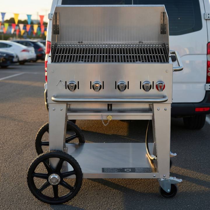 Crown Verity 30 inch mobile grill in stainless steel, windguard panels raised, parked on asphalt beside a white van