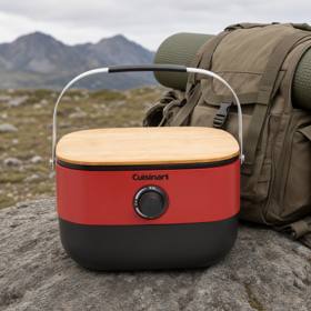Cuisinart Venture portable grill closed on a rock, red body with its wooden board on top and carry handle raised