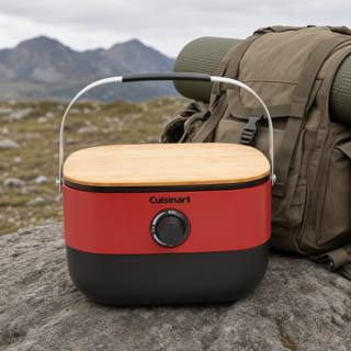 Cuisinart Venture portable grill closed on a rock, red body with its wooden board on top and carry handle raised