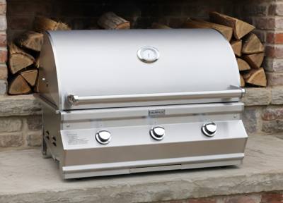 Fire Magic CM650i stainless grill head, hood closed with lid thermometer and three knobs, on stone before stacked firewood