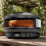 Gozney Arc gas pizza oven on a wooden table under a pergola, flame lit and dial showing 950