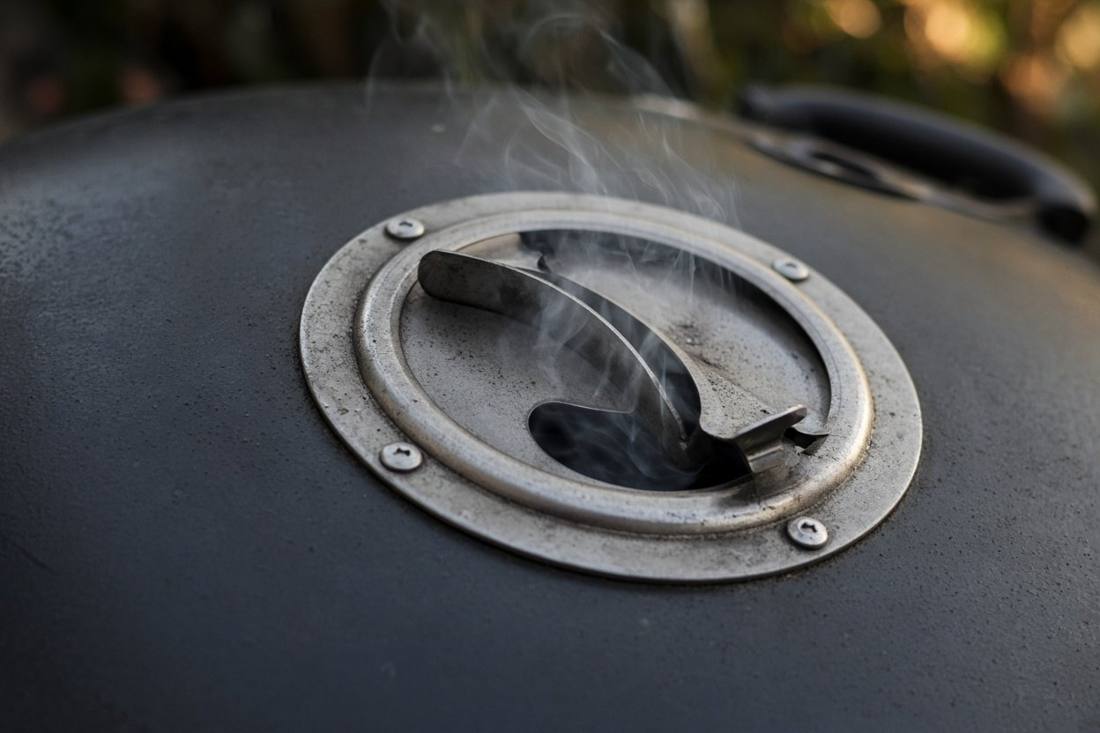 The metal damper vent on the lid of a black charcoal kettle grill, half open, with thin smoke escaping