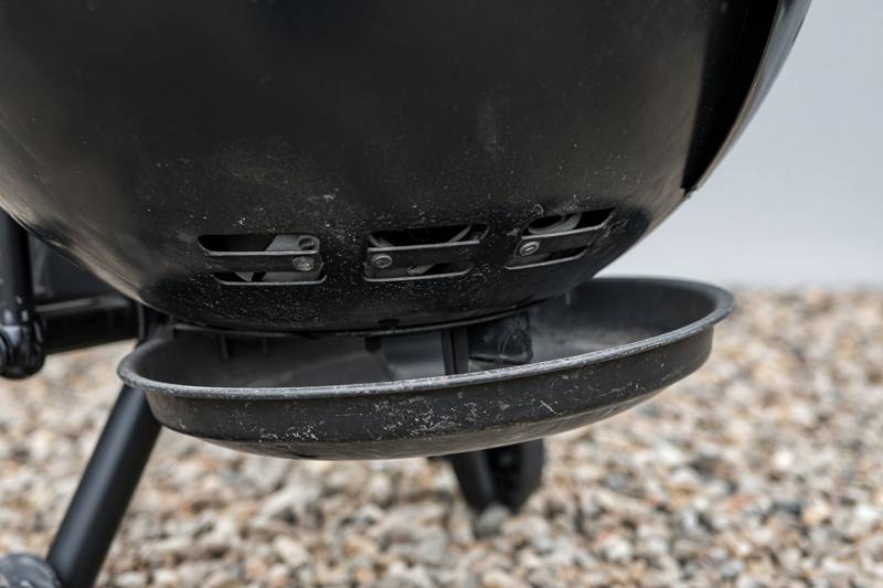 The bottom vent blades under the bowl of a black kettle grill, partly open, with the ash catcher beneath
