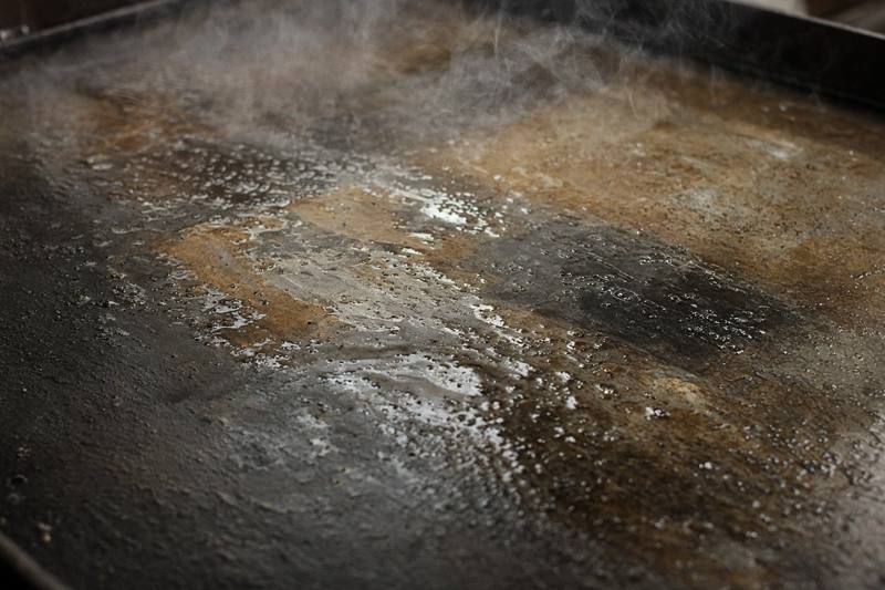 A hot steel griddle top with a thin film of oil, the steel turning bronze and brown in patches with some areas already black and light smoke rising