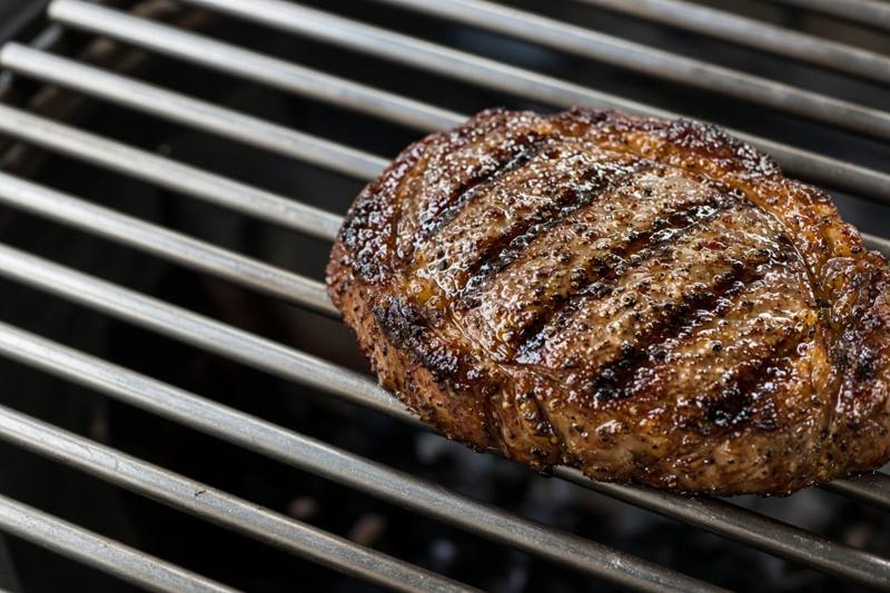 A ribeye steak with dark diagonal sear marks on thick, shiny stainless steel rod grates