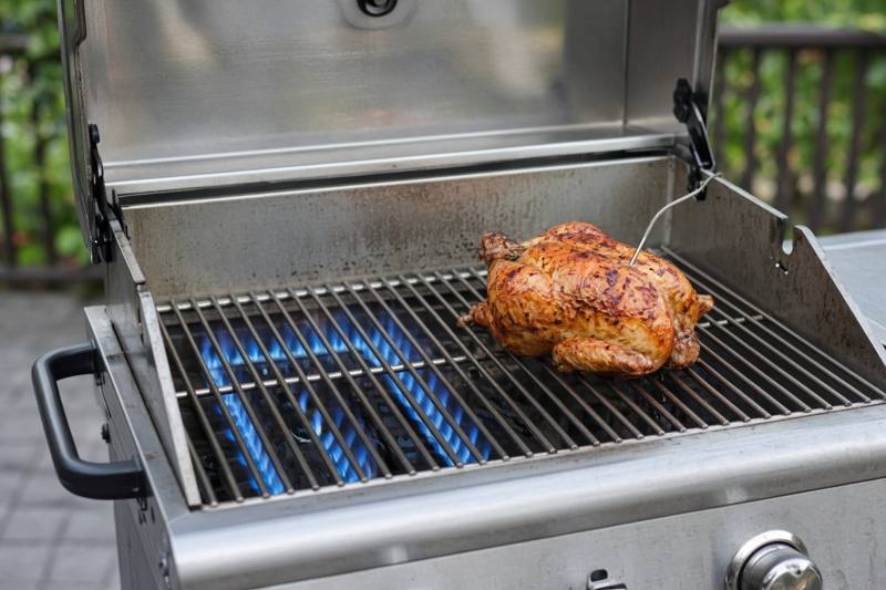 A stainless gas grill with blue flames under the left side of the grates and a whole chicken roasting on the unlit right side