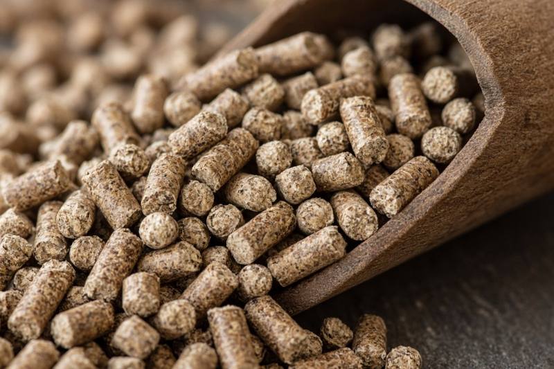 A pile of brown hardwood pellets, each a small cylinder about a quarter inch across, spilling from a wooden scoop