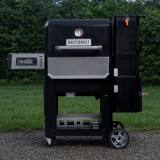 Masterbuilt Gravity Series 800 on gravel in front of a hedge, lid closed, hopper tower on the right