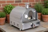 Mont Alpi stainless tabletop gas pizza oven on a stone counter beside potted herbs and a brick wall
