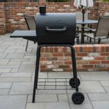 Black Nexgrill charcoal barrel grill with chimney and side shelf on gray paving beside a low brick wall