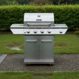 Stainless Nexgrill four-burner gas cart on wet gravel, rain-beaded lid, five knobs and two cabinet doors