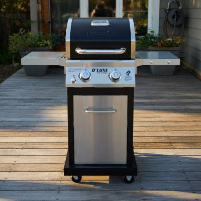 Nexgrill Deluxe two-burner gas grill on a timber deck, shelves out, stainless door shut, black lid closed.