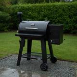 Black Nexgrill Oakford pellet barrel on a wet gravel pad by a hedge, front table down, controller lit