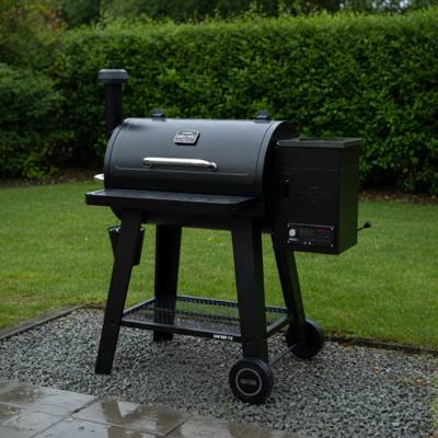 Black Nexgrill Oakford pellet barrel on a wet gravel pad by a hedge, front table down, controller lit