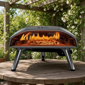 Ooni Koda 16 on a round wooden garden table under a pergola, flame lit with a pizza on the stone