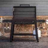 Traeger Irontop griddle against a stone wall, hood raised, tools hanging on the P.A.L. rails, propane tank below
