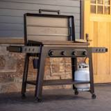 Traeger Irontop griddle on a porch beside a stone wall, lid closed, propane tank on the lower shelf