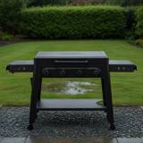 Black Traeger Irontop 4-burner griddle on wet gravel, lid closed, four control knobs and both side shelves out
