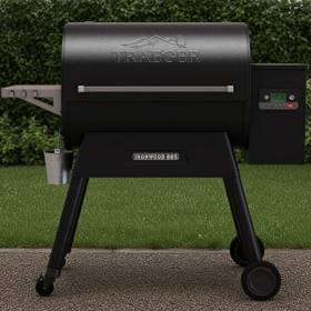 Black Traeger Ironwood 885 pellet grill on wet gravel beside a lawn, grease bucket hanging, clipped hedge behind