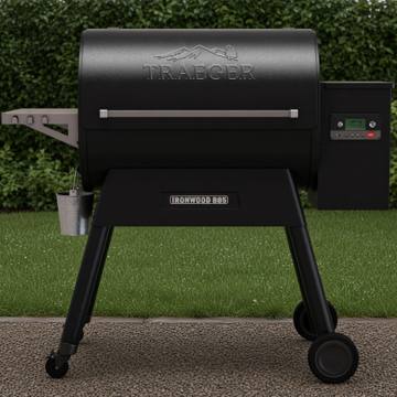 Black Traeger Ironwood 885 pellet grill on wet gravel beside a lawn, grease bucket hanging, clipped hedge behind