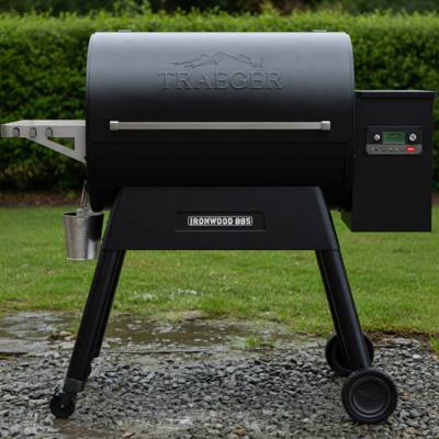 Black Traeger Ironwood 885 pellet grill on wet gravel beside a lawn, grease bucket hanging, clipped hedge behind