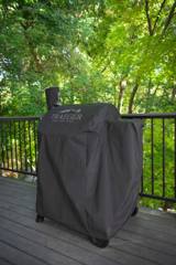 Black Traeger cover fitted over a pellet grill on a wooden deck with a metal railing and trees behind