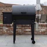 Black Traeger Tailgater pellet grill and grease bucket on grey paving, beside a fitted Traeger grill cover