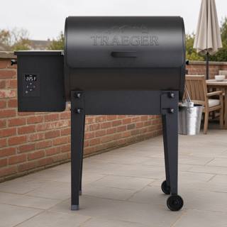 Black Traeger Tailgater pellet grill and grease bucket on grey paving, beside a fitted Traeger grill cover