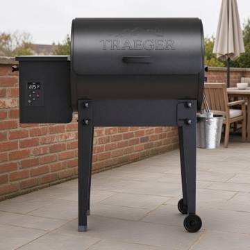 Black Traeger Tailgater pellet grill and grease bucket on grey paving, beside a fitted Traeger grill cover