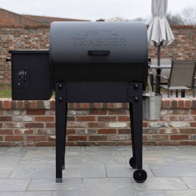 Black Traeger Tailgater pellet grill and grease bucket on grey paving, beside a fitted Traeger grill cover